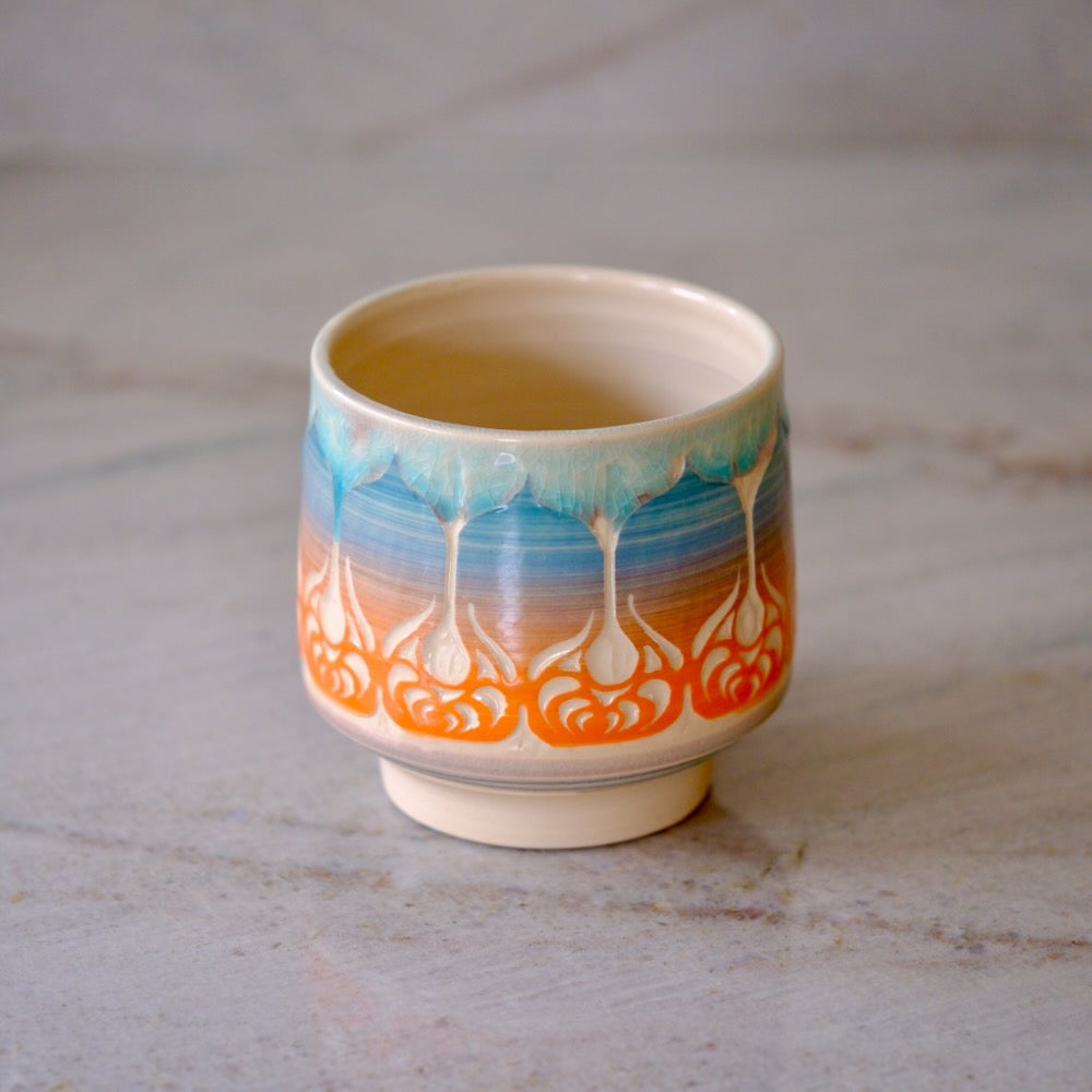 A small ceramic tea cup with a cream-colored glaze, decorated with a blue and orange wave-like pattern resembling stylized clouds or lotus motifs, set on a light gray marble surface.