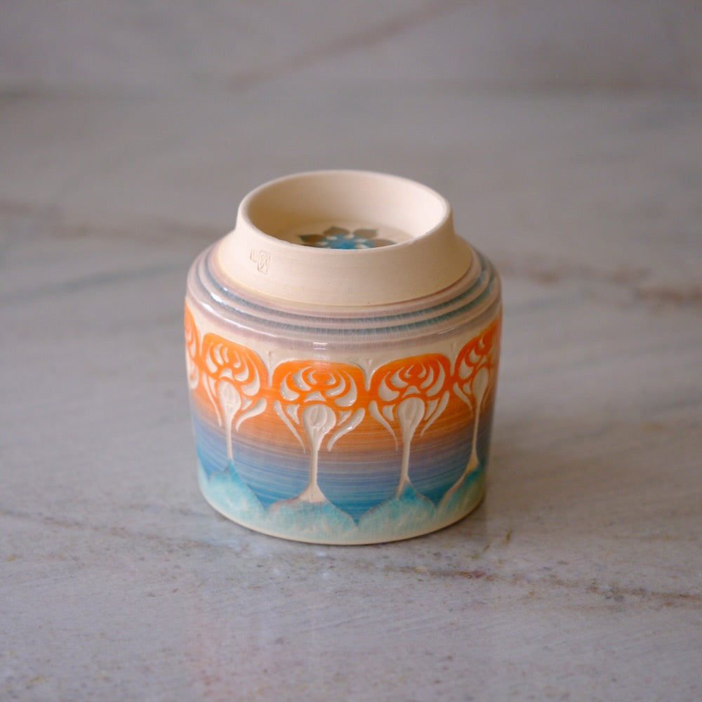 A small ceramic container with a lid, decorated with a colorful tree pattern in orange, yellow, blue, and white on a light gray marble surface.
