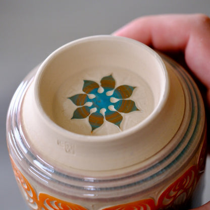 Close-up of a hand holding a ceramic jar, showing the interior lid with a painted blue and gold floral pattern on a beige ceramic surface.