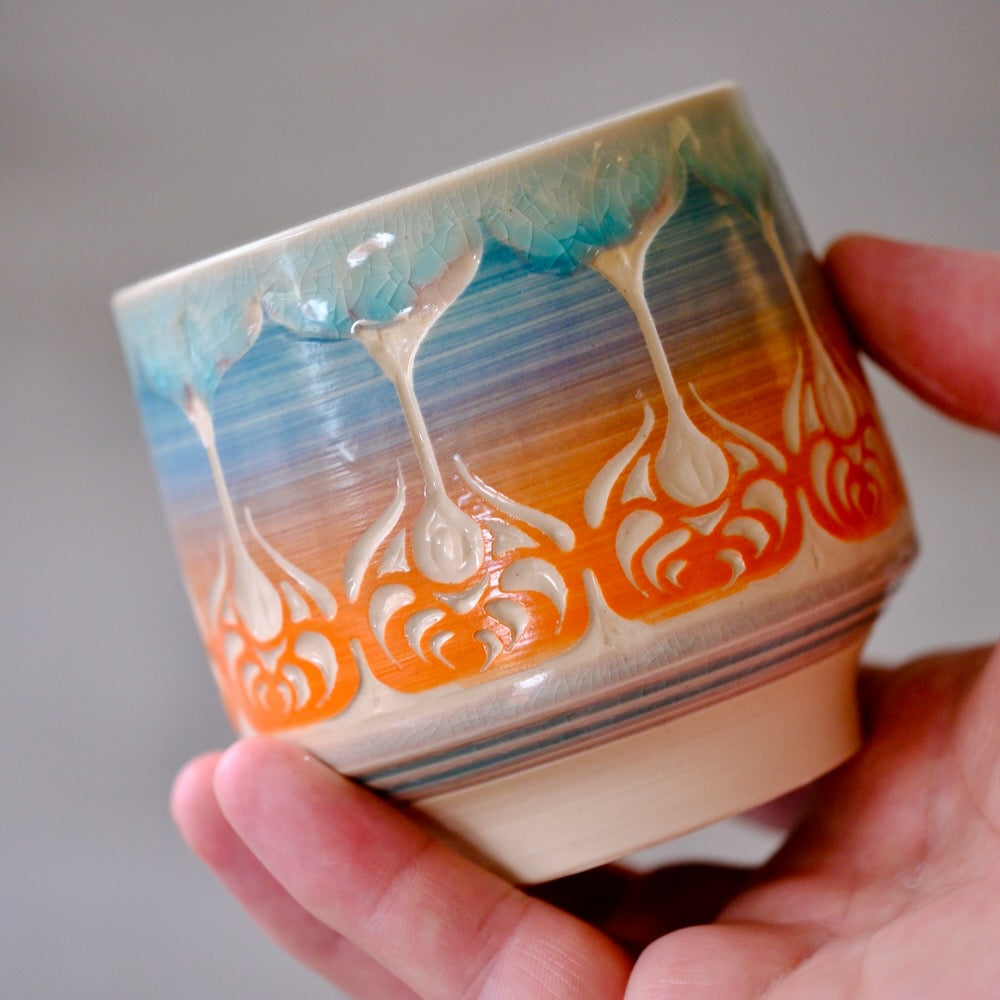A handmade ceramic tea cup with a blue, orange, and cream drip glaze pattern, held in a person's hand against a neutral gray background.