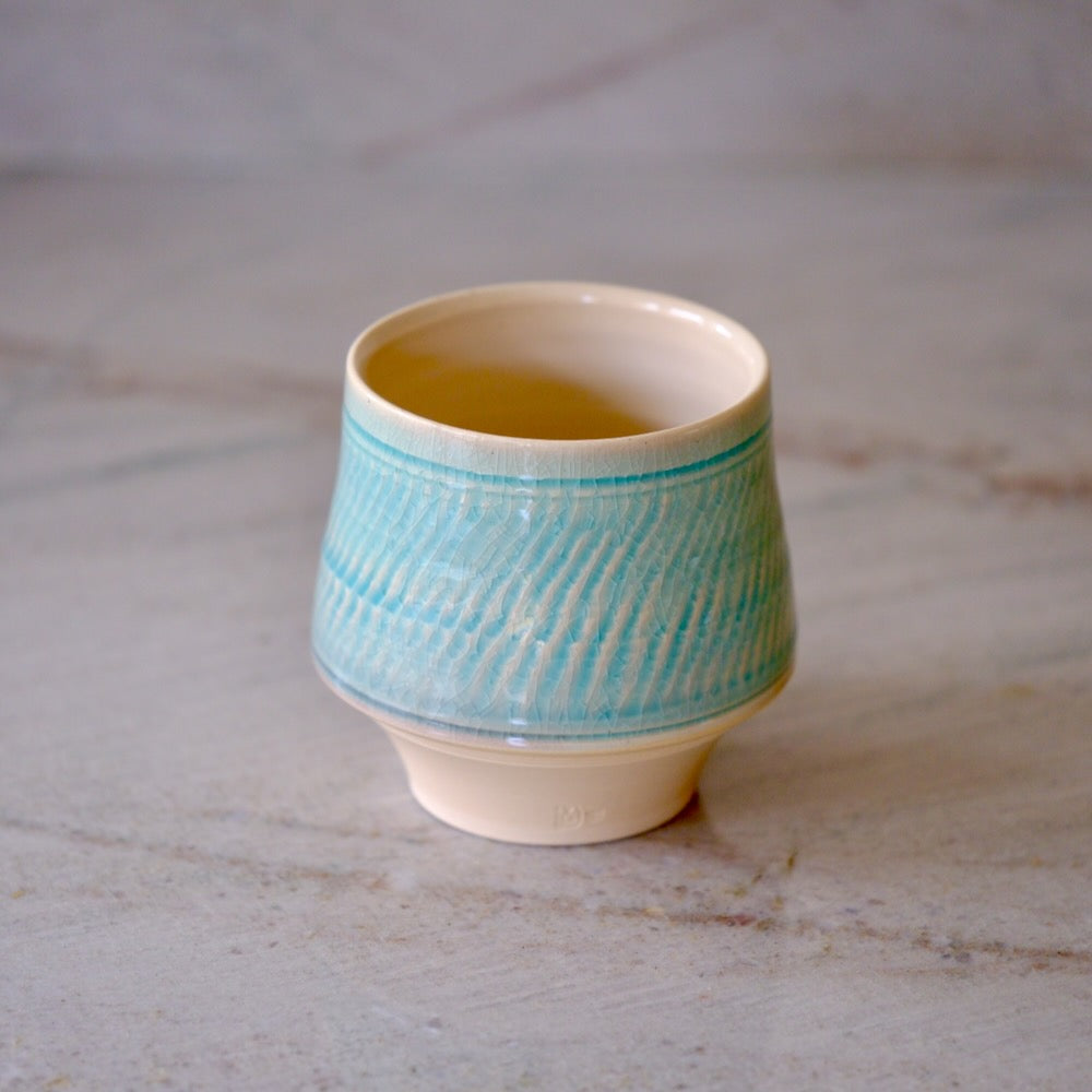 A small ceramic cup with a textured light blue and white patterned glaze, featuring a cream-colored interior and base, set on a light marble surface.