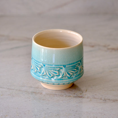 A small ceramic cup with a light blue-green glazed finish, featuring a turquoise pattern band around the middle and a warm cream interior, set on a light marble surface.