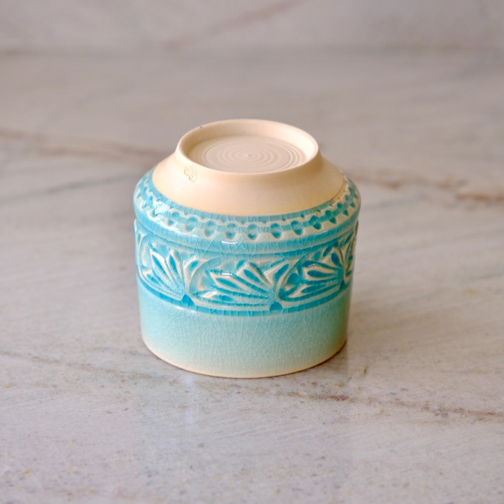 A small ceramic container or candle holder with a light blue and turquoise patterned glaze and a cream-colored interior, shown on a light marble surface.