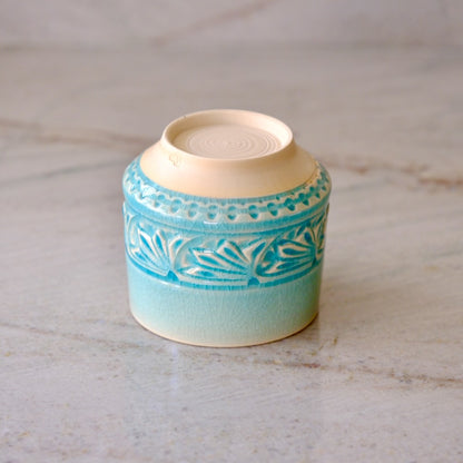 A small ceramic container or candle holder with a light blue and turquoise patterned glaze and a cream-colored interior, shown on a light marble surface.
