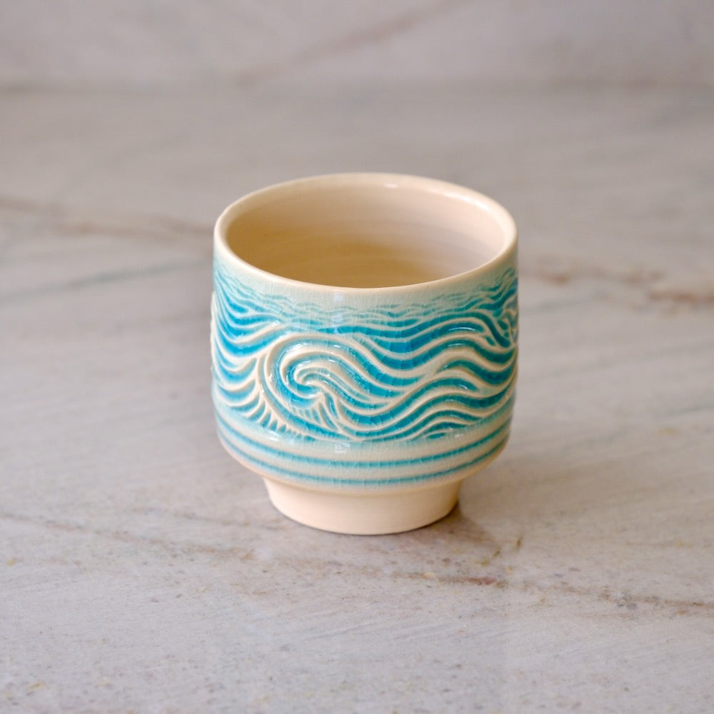 A small ceramic cup with a cream-colored interior and exterior base, decorated with a turquoise blue wave pattern around the middle, set on a light marble surface.