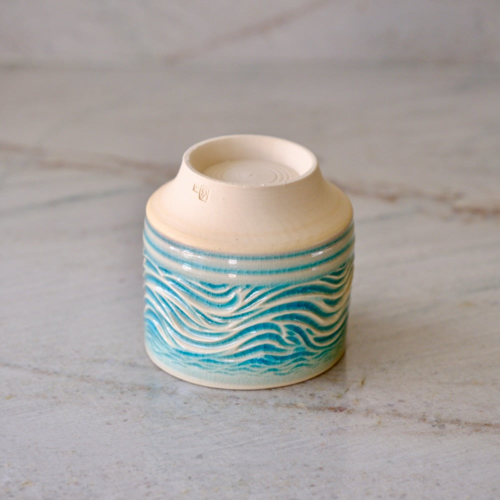 A small ceramic cup with a cream-colored interior and exterior base, decorated with a wavy turquoise and teal pattern on the outside, set on a light marble surface.