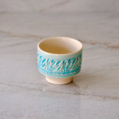 A small ceramic tea cup with a light beige interior and exterior base, decorated with a turquoise blue patterned band around the middle, set on a light marble surface.