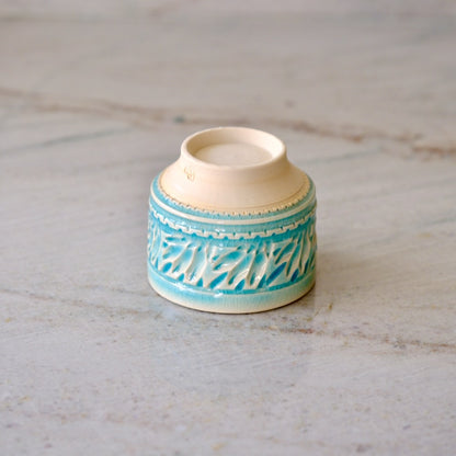 A small ceramic cup with a cream-colored interior and exterior base, decorated with a turquoise blue textured pattern around the lower half, set on a light stone surface.