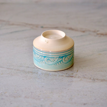 A small ceramic cup with a cream-colored top and a turquoise blue patterned lower half, shown on a light gray marble surface.
