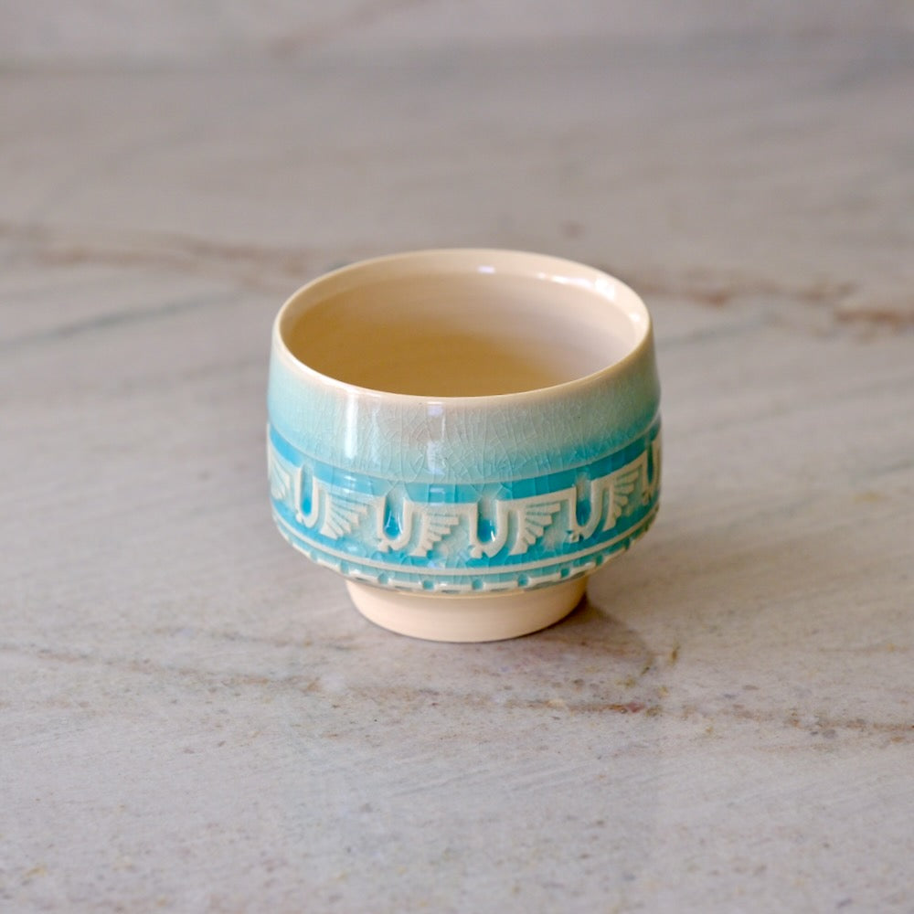 A small ceramic cup with a light beige body, a turquoise blue gradient upper section, and a decorative band of white Japanese characters around the middle, set on a light gray marble surface.