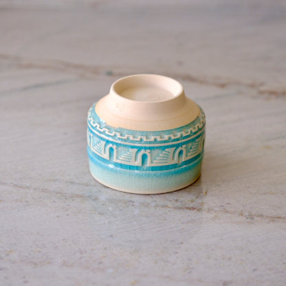 A small ceramic cup with a cream-colored interior and exterior, decorated with a turquoise blue patterned band around the middle, sitting on a light marble surface.
