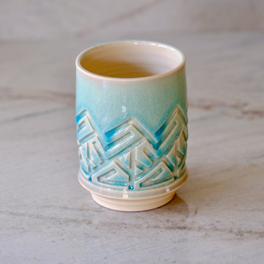 A small ceramic cup with a glossy light blue to cream gradient glaze and a raised geometric pattern around the middle, shown on a light marble surface.