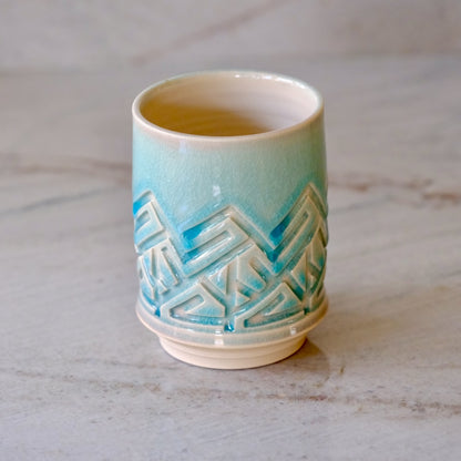 A small ceramic cup with a glossy light blue to cream gradient glaze and a raised geometric pattern around the middle, shown on a light marble surface.