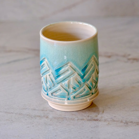 A small ceramic cup with a glossy light blue to cream gradient glaze and a raised geometric pattern around the middle, shown on a light marble surface.