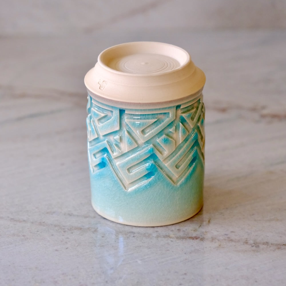 A ceramic cup with a cream-colored lid and a glazed body featuring a blue and white geometric pattern, set on a light marble surface.
