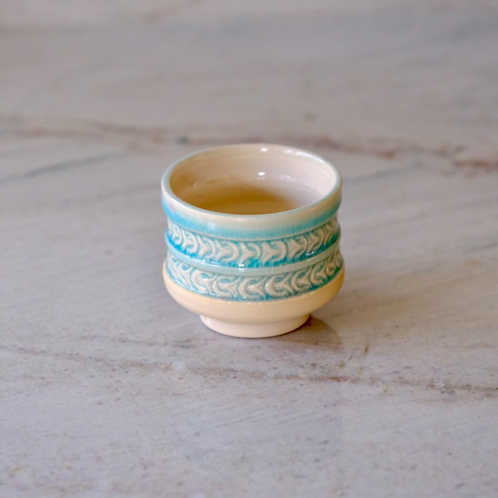 A small ceramic teacup with a cream-colored body and turquoise blue decorative bands, sitting on a light gray marble surface.