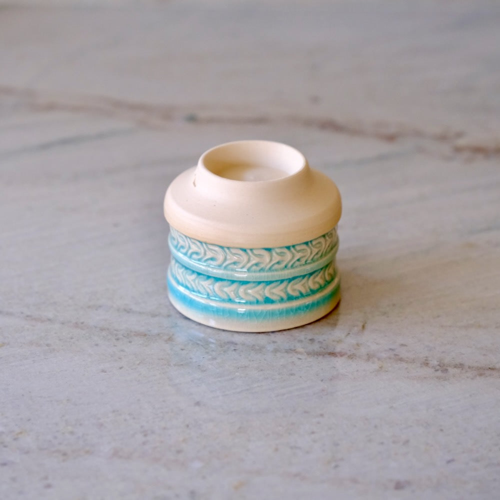 A small ceramic candle holder or candle tin with a cream-colored body and turquoise blue patterned bands, set on a light gray marble surface.