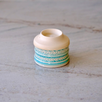 A small ceramic candle holder or candle tin with a cream-colored body and turquoise blue patterned bands, set on a light gray marble surface.