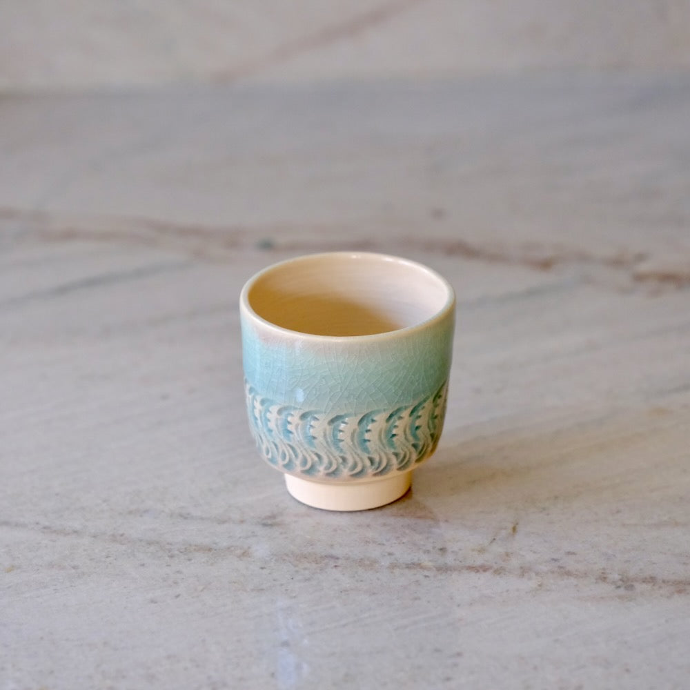 A small ceramic sake cup with a light blue-green glazed body, a textured wave pattern, and a cream-colored interior, set on a light gray marble surface.
