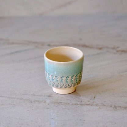 A small ceramic sake cup with a light blue-green glazed body, a textured wave pattern, and a cream-colored interior, set on a light gray marble surface.