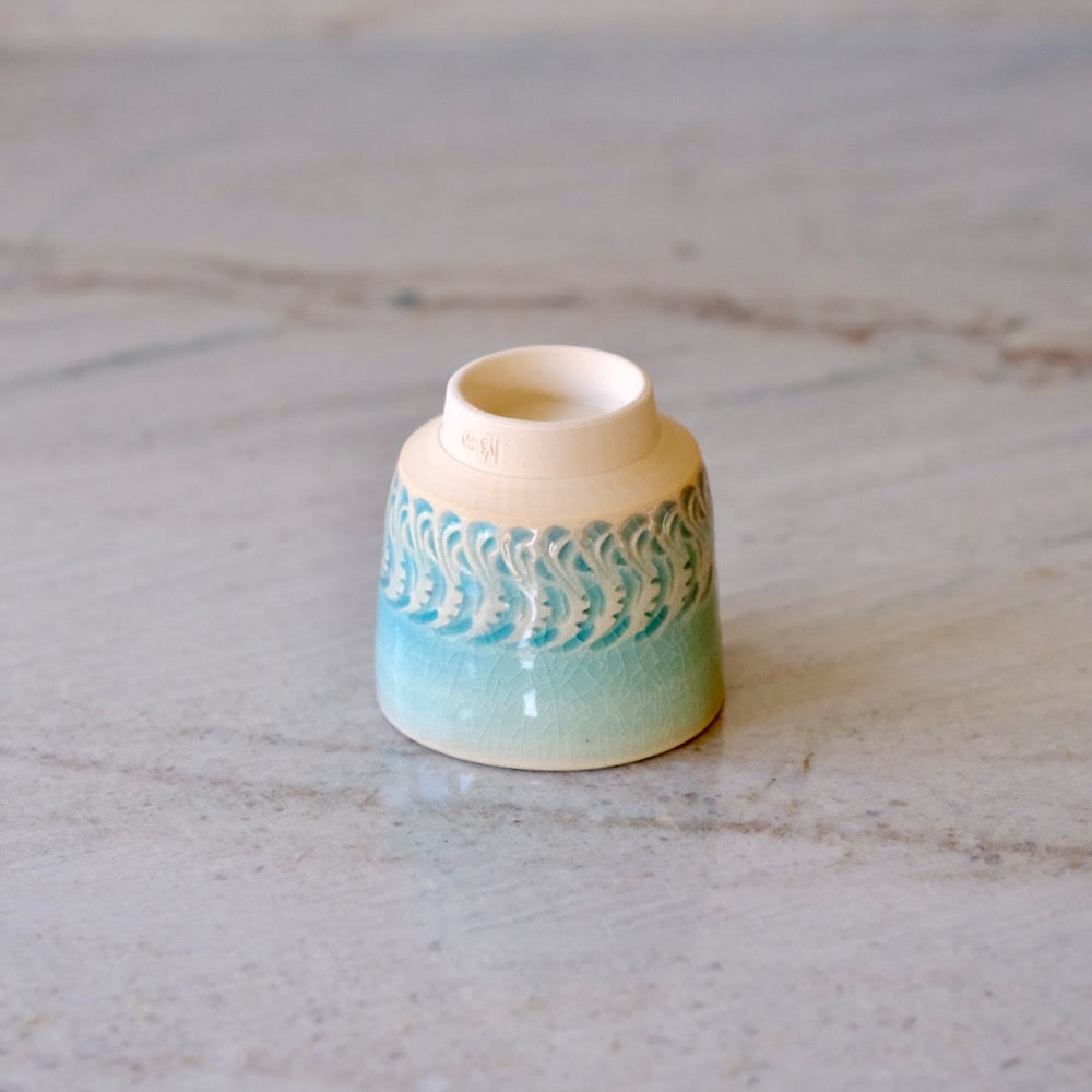 A small ceramic cup with a cream-colored interior and exterior, featuring a turquoise and light blue wave pattern around the middle, resting on a light marble surface.