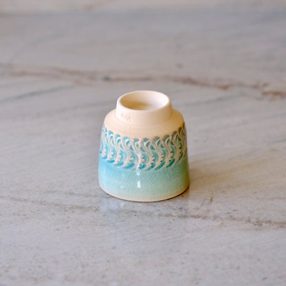 A small ceramic cup with a cream-colored interior and exterior, featuring a turquoise and light blue wave pattern around the middle, resting on a light marble surface.