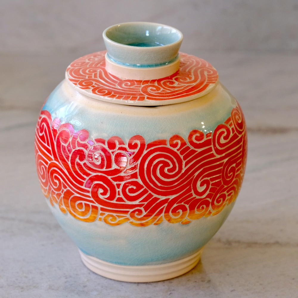 A glazed ceramic lidded vase with a rounded body, featuring a light blue-green glaze and a bold red-orange scroll pattern around the middle, set against a neutral gray background.