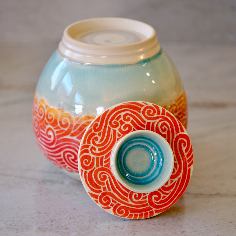 A glazed ceramic jar with a matching lid placed in front. The jar has a pale aqua upper body, orange and red wave-like patterns around the lower half, and a cream-colored interior and lid base.