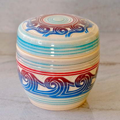 A round ceramic lidded container with a cream-colored glaze, decorated with blue and red wave-like patterns and horizontal blue stripes, set on a light marble surface.