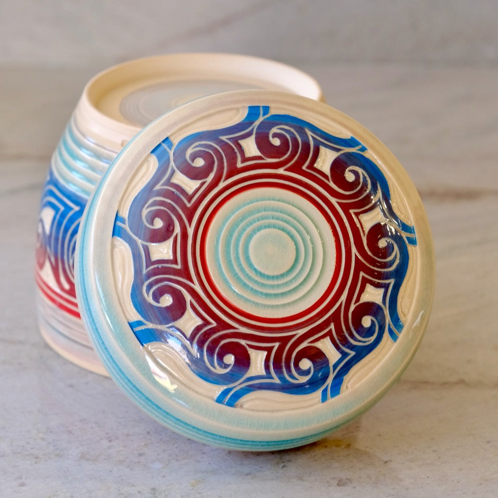 A ceramic lidded container with a round lid featuring a colorful circular mandala pattern in blue, red, and light blue on an off-white base, with the matching container partially visible behind it on a light stone surface.