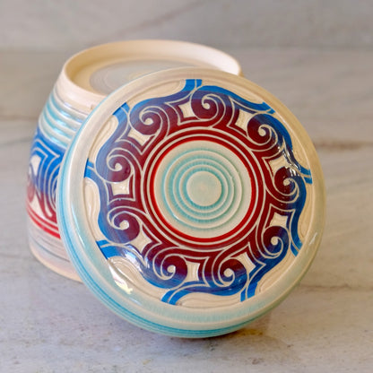 A ceramic lidded container with a round lid featuring a colorful circular mandala pattern in blue, red, and light blue on an off-white base, with the matching container partially visible behind it on a light stone surface.