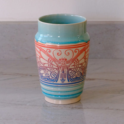 A ceramic vase with a light blue-green glazed finish, featuring a central cream-colored band decorated with a red and blue butterfly design, set on a light gray marble surface.