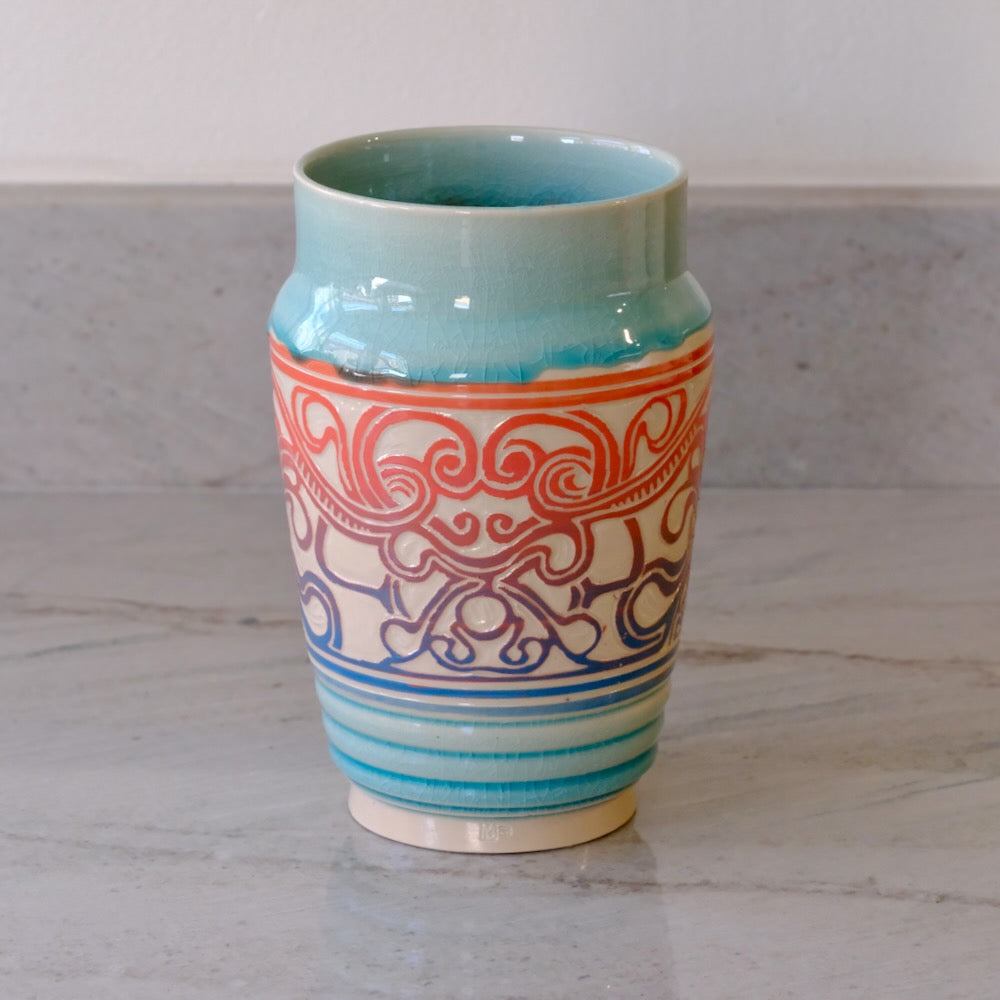 A ceramic vase with a glossy light blue-green glazed finish, featuring a cream-colored middle section decorated with red and blue scrollwork patterns, and a striped base.
