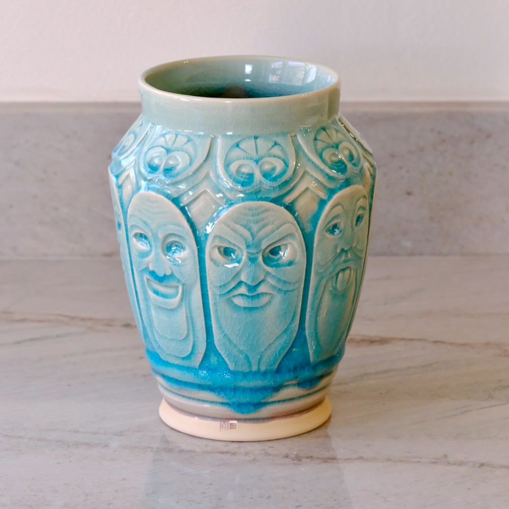 A ceramic vase with a glossy turquoise blue glaze and raised relief carvings of three faces, sitting on a light marble countertop.