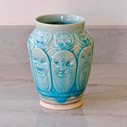 A ceramic vase with a glossy turquoise blue glaze and raised relief carvings of three faces, sitting on a light marble countertop.