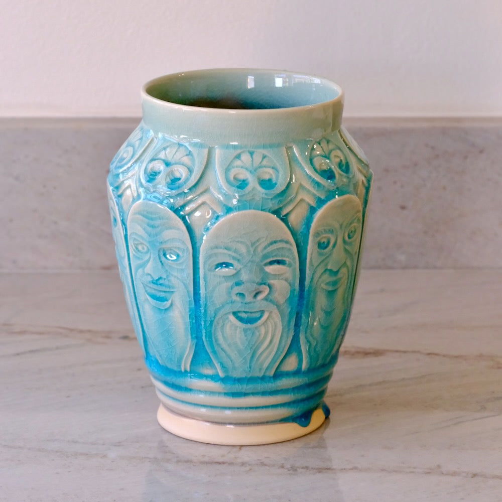 A ceramic vase with a glossy turquoise blue glaze and raised relief faces, standing on a light marble surface against a neutral background.