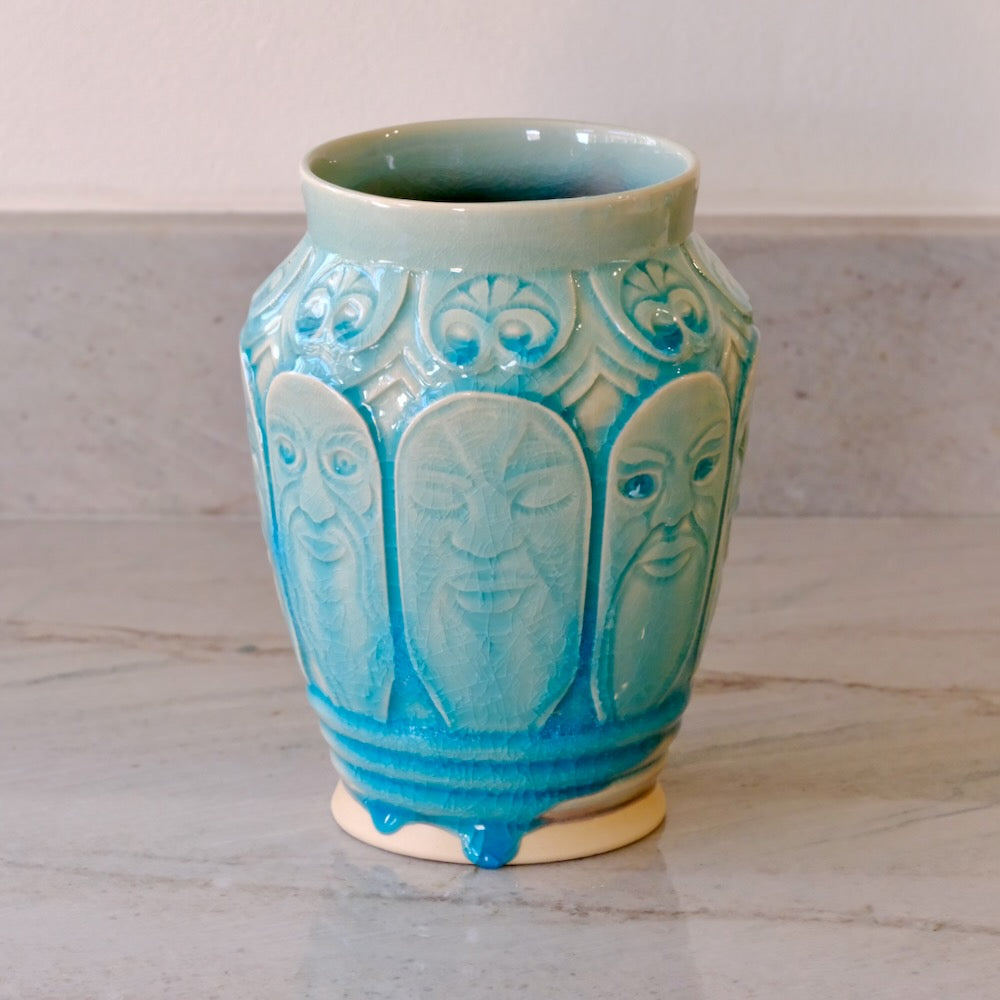 A ceramic vase with a glossy turquoise blue glaze, featuring embossed stylized faces and floral motifs, set on a light gray marble surface against a neutral background.